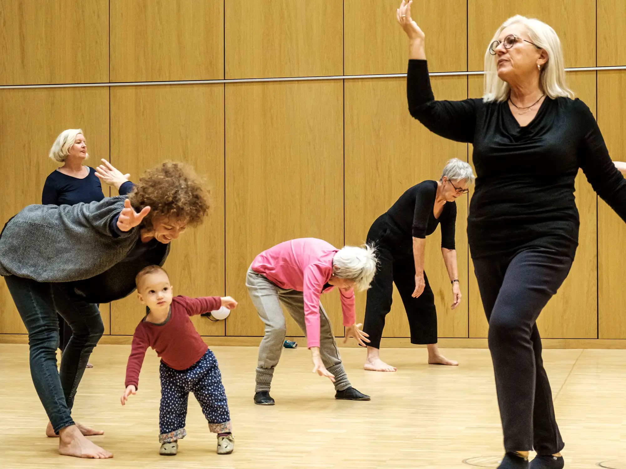 Menschen verschiedenen Alters bewegen sich in einem Raum. Eine Frau tanzt mit einem kleinen Kind.