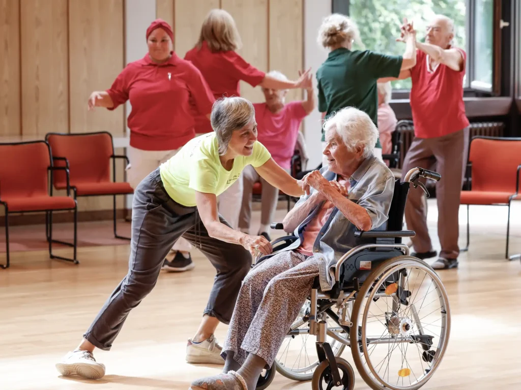 Ältere Menschen tanzen gemeinsam in einem Raum. Im Vordergrund tanzen eine Tanzvermittlerin und eine Frau im Rollstuhl.