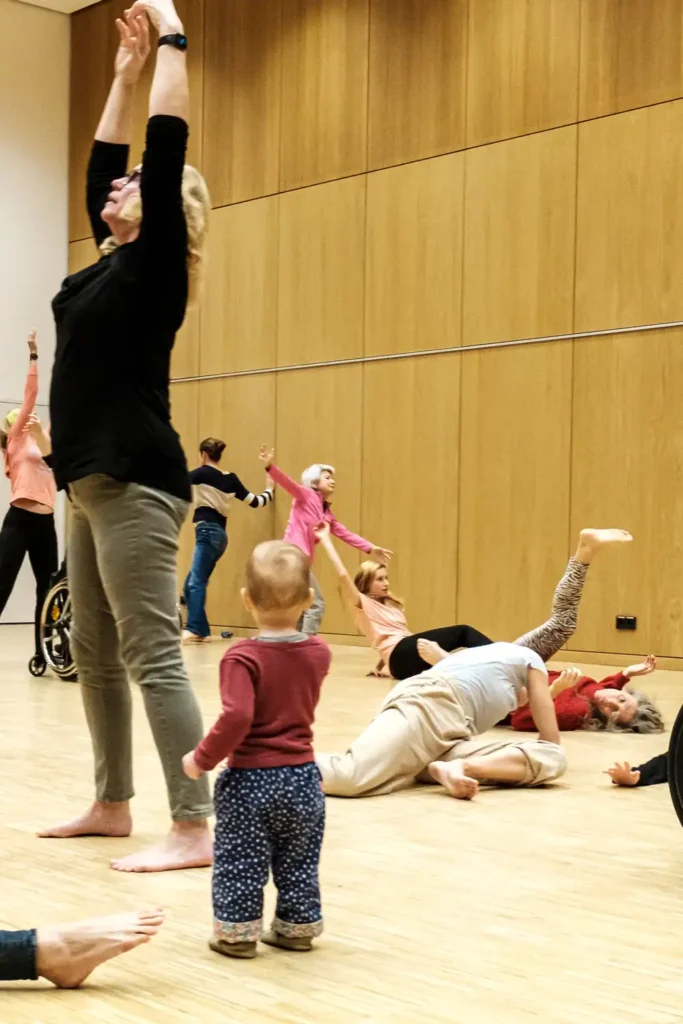 Im Vordergrund steht ein kleines Kind und blickt auf eine Gruppe tanzender Menschen. Sie bewegen sich zum Teil am Boden.