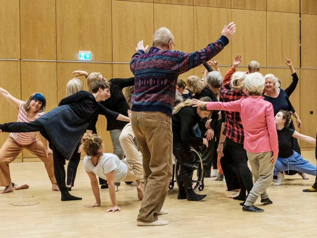 Viele Menschen tanzen zusammen. Eine Frau bewegt sich am Boden. Ein Mann ist von hinten zu sehen, seine Arme sind nach oben ausgebreitet.
