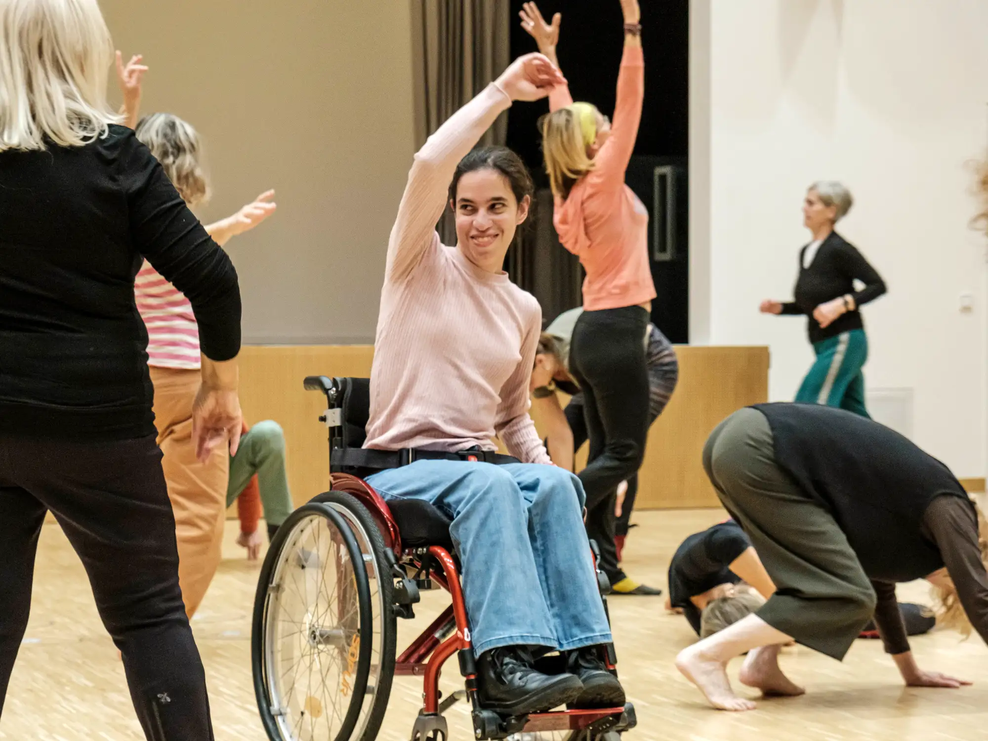 Eine junge Frau im Rollstuhl tanzt mit gehobenem Arm. dahinter bewgt sich eine andere Person mit erhobenen Armen.