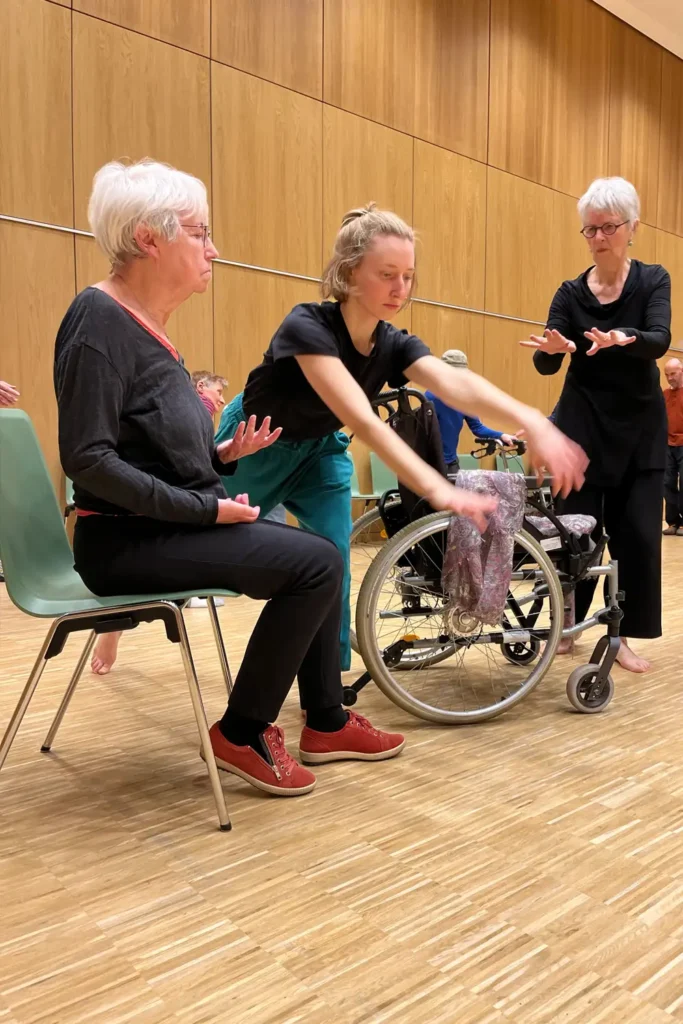 Drei Frauen tanzen mit ihren Händen. Eine der Frauen sitzt auf einem Stuhl. 